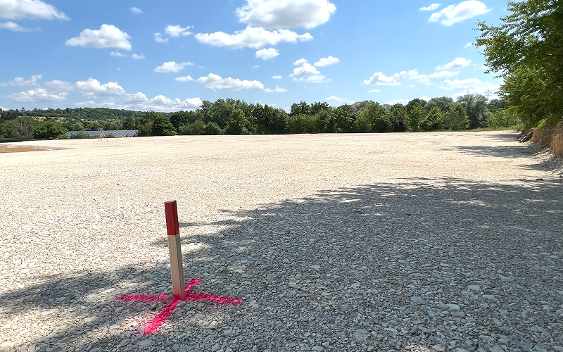 Ein Holzpfahl mit rotem oberen Ende und pinker Markierung auf einem Kiesgrundstück.