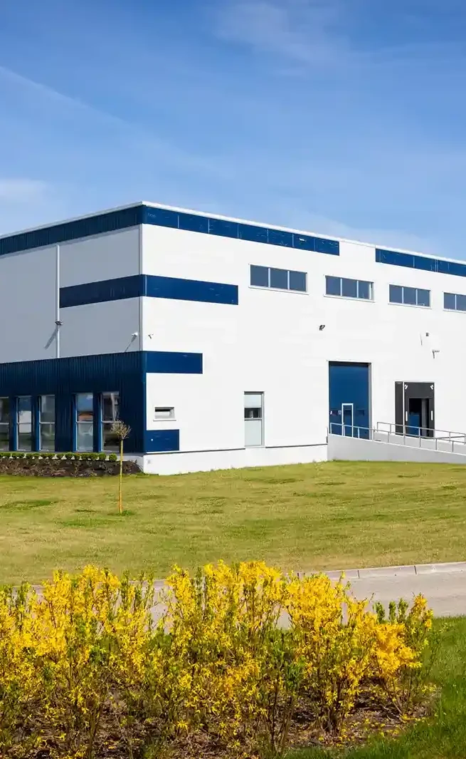 Moderne Lagerhalle mit blauer und weißer Fassade, umgeben von Rasen und gelben Blumen.