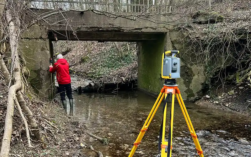 Ein Vermessungstechniker führt Messungen an einer Brücke in einem Waldgebiet durch, während ein Vermessungsgerät im Vordergrund steht.