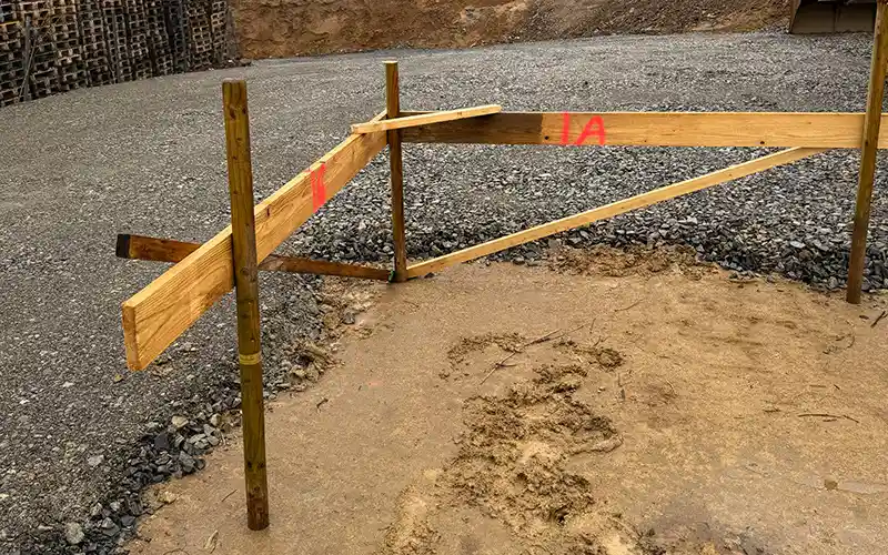 Holzrahmen mit roten Markierungen auf einer Baustelle, umgeben von Kies und Sand.